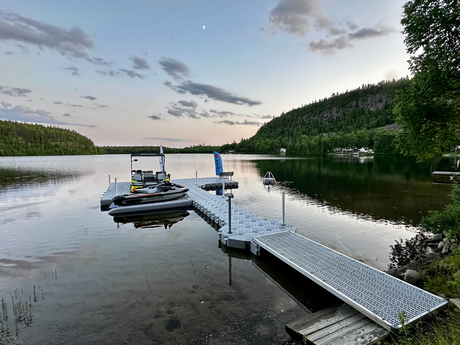 A floating dock with a moored jet ski extends into a calm lake surrounded by forested hills at dusk.