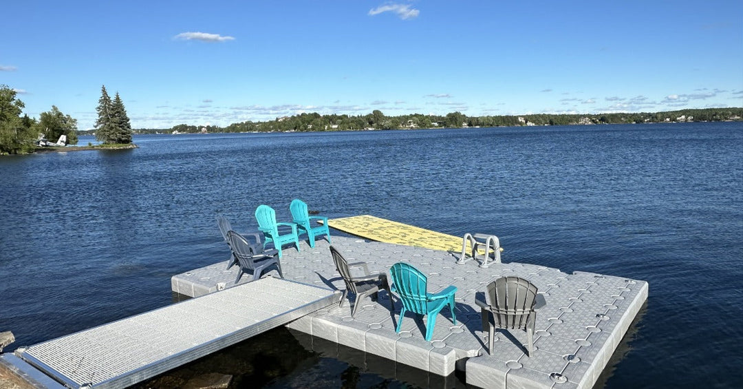 A modular floating dock with chairs sits on a calm lake, connected to shore by a ramp under a clear blue sky.